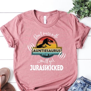 May include: A pink t-shirt with a white graphic design that says "Don't mess with Auntieosaurus you'll get Jurasskicked". The graphic features a dinosaur in a retro sunset design.