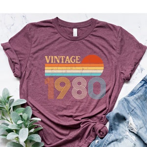May include: A maroon t-shirt with a vintage 1980 graphic design featuring a sunset and rainbow stripes. The graphic is in shades of orange, yellow, blue, and green.
