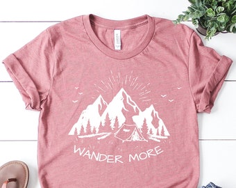 Könnte beinhalten: Ein staubiges rosafarbenes T-Shirt mit einer weißen Grafik von Bergen und einem Zelt mit dem Text "Wander More".