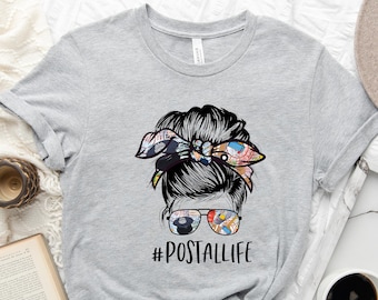 May include: A gray t-shirt with a graphic of a woman with a messy bun and sunglasses. The bun is decorated with a colorful bandana. The text "#POSTALLIFE" is printed below the graphic.
