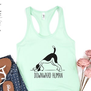 May include: A light green tank top with a black and white illustration of a dog doing a downward facing dog yoga pose. The text "Downward Human" is printed below the dog.