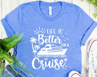 May include: A blue t-shirt with a white graphic that says "Life is Better on a Cruise" with a cruise ship and palm trees. The shirt is on a white wooden surface with a pair of white sneakers and blue denim shorts.
