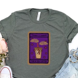 Puede incluir: Una camiseta verde con un dise&ntilde;o de carta de tarot que presenta un caf&eacute; helado con una pajita y el texto "The Iced Coffee". La camiseta se combina con pantalones cortos de mezclilla azul claro y zapatillas blancas.