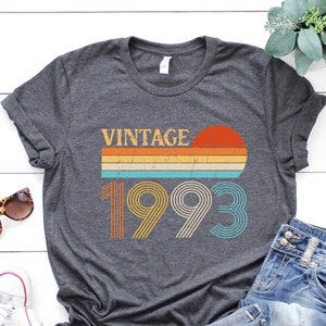 May include: A dark grey t-shirt with a vintage 1993 design. The design features a sunset with a rainbow stripe and the year 1993 in a retro font.