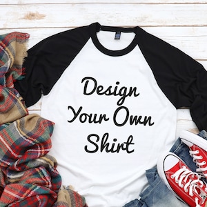 May include: A white and black baseball tee shirt with the text "Design Your Own Shirt" on the front. The shirt is laying on a white wooden surface with a plaid scarf, a red ornament, and a pair of red sneakers.
