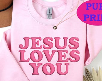 May include: A light pink sweatshirt with the phrase "JESUS LOVES YOU" in a raised, bubble-style font. The words are in a darker shade of pink. A gold necklace and a brown hat are also visible.