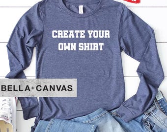 May include: A heather blue long-sleeve shirt with the white text CREATE YOUR OWN SHIRT printed on the chest. The shirt is displayed with a pair of blue denim jeans and a single white canvas sneaker with a rubber sole and white laces.