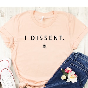 May include: A peach-colored t-shirt with the text "I DISSENT." in black lettering and a small crown with the letters "RBG" underneath. The shirt is laid out on a white wooden surface with a pair of white sneakers and blue jeans.