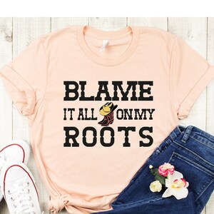 May include: A peach-colored t-shirt with the text "Blame it all on my roots" in black lettering. A cartoon boot with a cowboy hat is in the middle of the text. The shirt is laid out on a wooden surface with a pair of white sneakers and blue jeans.