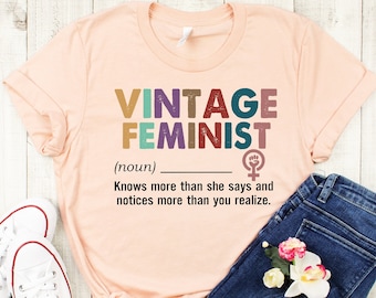May include: A peach-colored t-shirt with the text "VINTAGE FEMINIST (noun) Knows more than she says and notices more than you realize." in colorful letters. The text is in a vintage style font. A feminist symbol is next to the text.