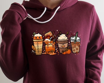 May include: A maroon hoodie featuring a graphic of four autumn-themed coffee drinks with pumpkin spice, whipped cream, and sprinkles. The drinks are set against a backdrop of autumn leaves and pumpkins.