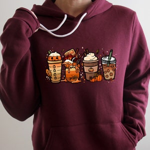 May include: A maroon hoodie with a graphic of four fall-themed coffee drinks with pumpkin spice, whipped cream, and sprinkles. The drinks are in front of a background of fall leaves and pumpkins.