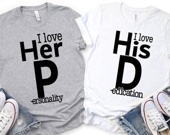 May include: Two gray and white t-shirts with black text. The gray shirt says "I love Her P-ersonality" and the white shirt says "I love His D-education".