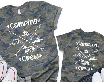 Könnte beinhalten: Zwei Camouflage-T-Shirts mit dem Text "Camping Crew" und einer Camping-Grafik. Die Shirts liegen auf einem weißen Hintergrund mit Jeans und Turnschuhen.