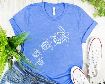 May include: A blue t-shirt with a white outline of four sea turtles of different sizes. The turtles are facing the right side of the image. The t-shirt is laying on a wooden surface with a pair of white sneakers and a pair of blue denim shorts.