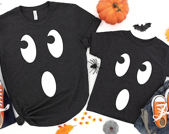 May include: Two black t-shirts with a white ghost face design. The larger shirt has two eyes and a mouth, while the smaller shirt has two eyes and a mouth. The shirts are laid out on a white background with autumn decorations.
