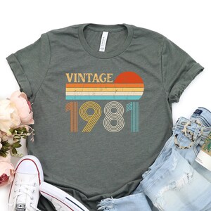 May include: A dark green t-shirt with a vintage 1981 design. The design features a sunset with a rainbow stripe and the year 1981 in a retro font.