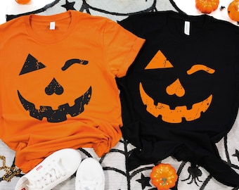 May include: Two t-shirts with a winking jack-o'-lantern face design. One shirt is orange with a black jack-o'-lantern face and the other is black with an orange jack-o'-lantern face.