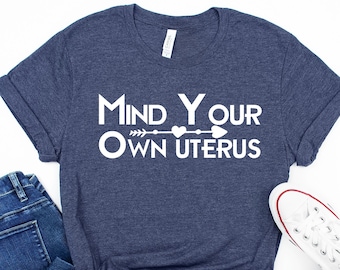 Könnte beinhalten: Ein marineblaues T-Shirt mit dem Text "Mind Your Own Uterus" in Weiß mit einem Herz- und Pfeil-Motiv. Das Shirt liegt flach auf einem weißen Hintergrund mit einem Paar weißen Canvas-Schuhen und einer blauen Jeans.