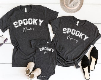 May include: Three dark grey t-shirts with the text "Spooky" in white, with the words "Daddy", "Mommy", and "Mom" underneath. The shirts are on a white wooden surface.