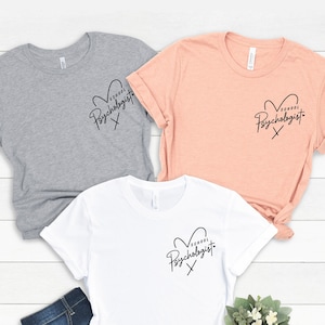 May include: Three t-shirts in different colors, gray, peach, and white, with the text "School Psychologist" in a heart shape design.