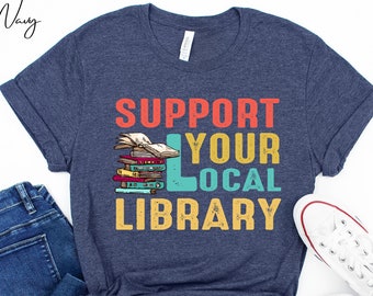 May include: Navy blue t-shirt with the text "SUPPORT YOUR LOCAL LIBRARY" in red, teal, yellow, and orange. The graphic includes a stack of books. A pair of blue jeans and white sneakers are also in the image.