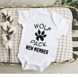 May include: White baby onesie with black text that reads "Wolf Pack New Member" and a black paw print with a small house inside.