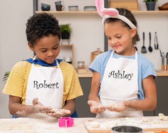 Könnte beinhalten: Zwei Kinder tragen weiße Schürzen mit ihren Namen, Robert und Ashley, darauf gestickt, und backen in der Küche. Beide sind mit Mehl bedeckt und haben einen verspielten Gesichtsausdruck.