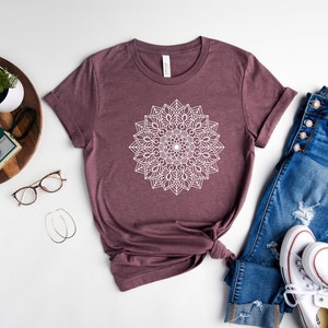 May include: A mauve t-shirt with a white mandala design. The t-shirt is laid flat on a white surface with a pair of jeans and white sneakers.
