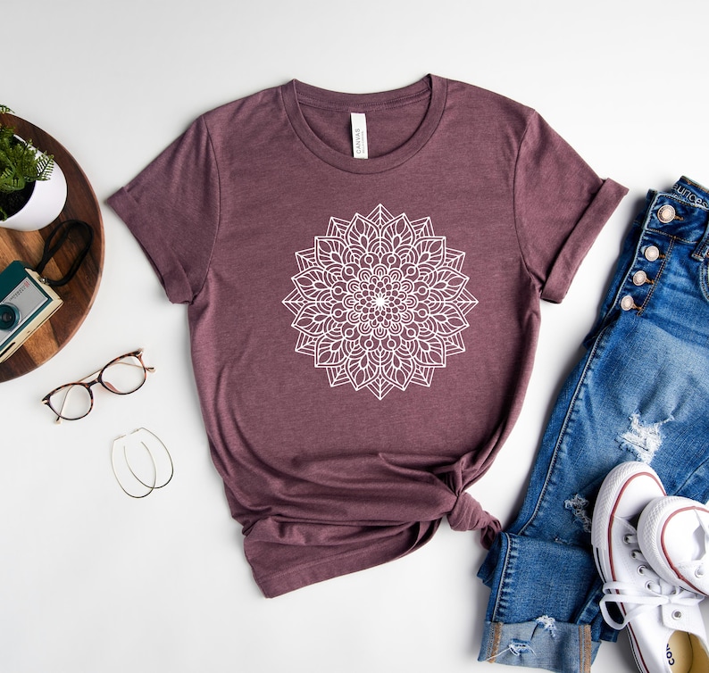 May include: A mauve t-shirt with a white mandala design. The t-shirt is laid flat on a white surface with a pair of jeans and white sneakers.