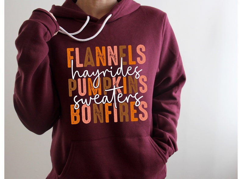 May include: A burgundy hoodie with a white drawstring and a front pocket. The front of the hoodie displays the words "FLANNELS, hayrides, PUMPKINS, sweaters, BONFIRES" in orange, brown, and white, suggesting an autumn theme.