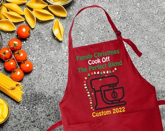 Op de afbeelding: Rode kerst-schort met de tekst "Family Christmas Cook Off The Perfect Blend" en een zwart-witte illustratie van een standmixer met kerstverlichting. De tekst "Custom 2022" staat onder de illustratie.