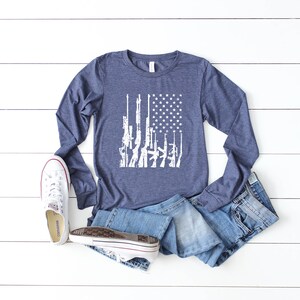 May include: A navy blue long-sleeved t-shirt with a white graphic of an American flag made up of guns. The shirt is laid out on a white wooden surface with a pair of white sneakers and blue jeans.