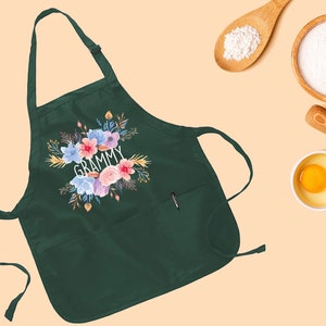 May include: A dark green apron with a floral design and the word "GRAMMY" in white lettering. The apron has a pocket on the front and a tie strap.