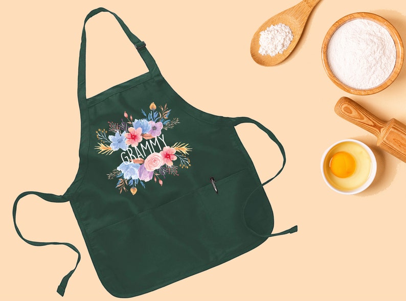 May include: A dark green apron with a floral design and the word "GRAMMY" in white lettering. The apron has a pocket on the front and a tie strap.
