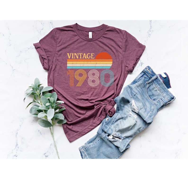 May include: A maroon t-shirt with a vintage 1980 graphic design featuring a sunset and rainbow stripes. The graphic is in shades of orange, yellow, blue, and green.