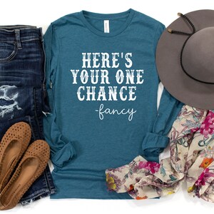 May include: Teal long-sleeve shirt with the text "Here's your one chance -fancy" in white. The shirt is paired with blue jeans, brown shoes, a floral scarf, and a gray hat.