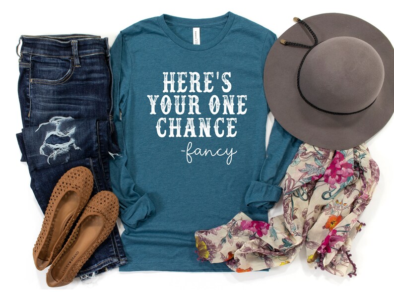 May include: Teal long-sleeve shirt with the text "Here's your one chance -fancy" in white. The shirt is paired with blue jeans, brown shoes, a floral scarf, and a gray hat.