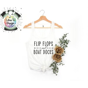 May include: White tank top with the text "Flip Flops and Boat Docks" printed on it. The tank top is hanging on a wooden hanger with a sprig of greenery and two brown fabric flowers.