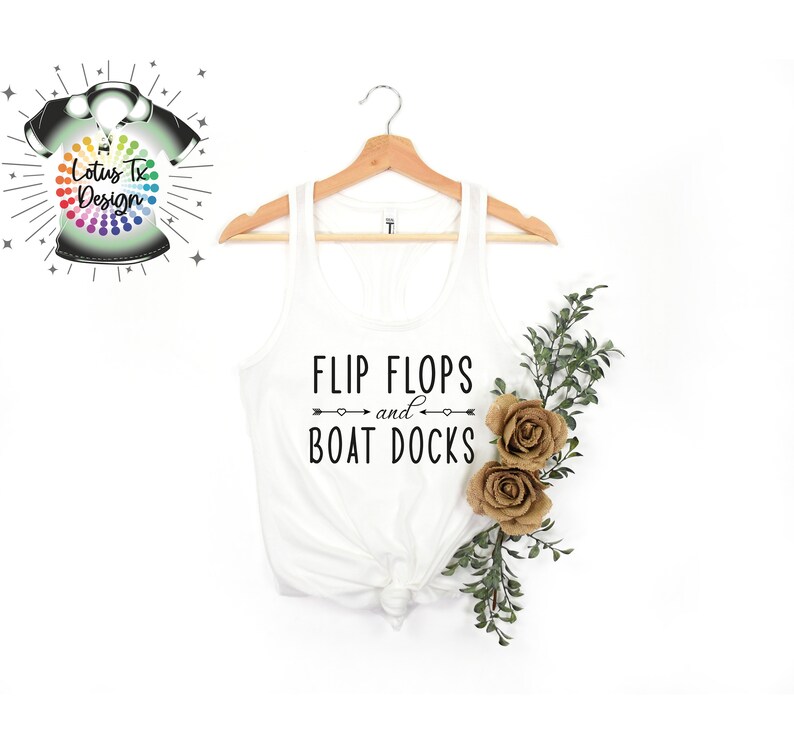 May include: White tank top with the text "Flip Flops and Boat Docks" printed on it. The tank top is hanging on a wooden hanger with a sprig of greenery and two brown fabric flowers.