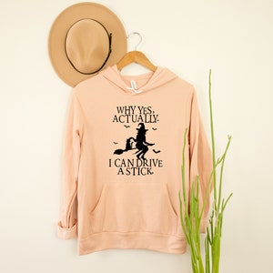 May include: A peach-colored hooded sweatshirt with a black graphic of a witch riding a broomstick with a black cat. The text on the sweatshirt reads "Why yes, actually. I can drive a stick."