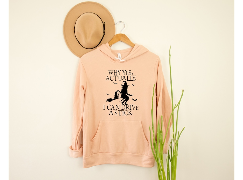 May include: A peach-colored hooded sweatshirt with a black graphic of a witch riding a broomstick with a black cat. The text on the sweatshirt reads "Why yes, actually. I can drive a stick."