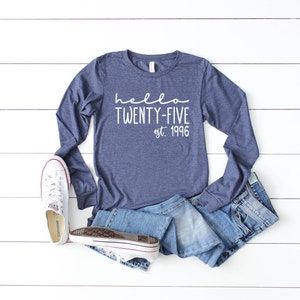 May include: A navy blue long-sleeved shirt with the text "hello twenty-five est. 1996" in white. The shirt is laid out on a white wooden surface with a pair of white sneakers and blue jeans.