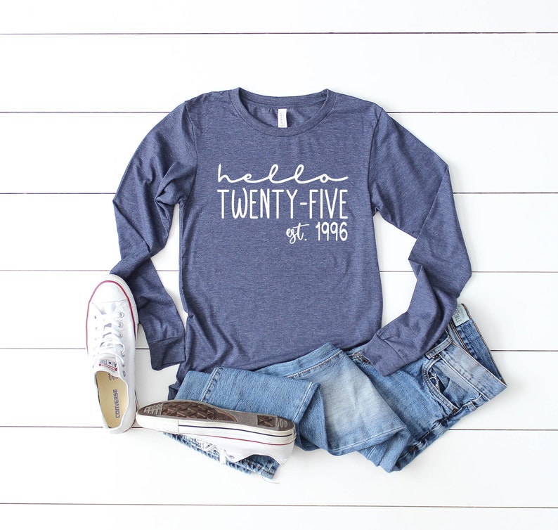May include: A navy blue long-sleeved shirt with the text "hello twenty-five est. 1996" in white. The shirt is laid out on a white wooden surface with a pair of white sneakers and blue jeans.