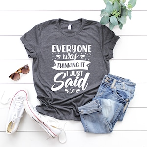 May include: A dark gray t-shirt with the text "Everyone was thinking it I just said it" in white. The shirt is laid out on a white wooden surface with a pair of sunglasses, a pair of white sneakers, and a pair of blue jeans.