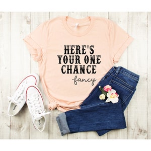 May include: A peach-colored t-shirt with the text "HERE'S YOUR ONE CHANCE -fancy" printed in black. The shirt is paired with blue jeans and white sneakers.