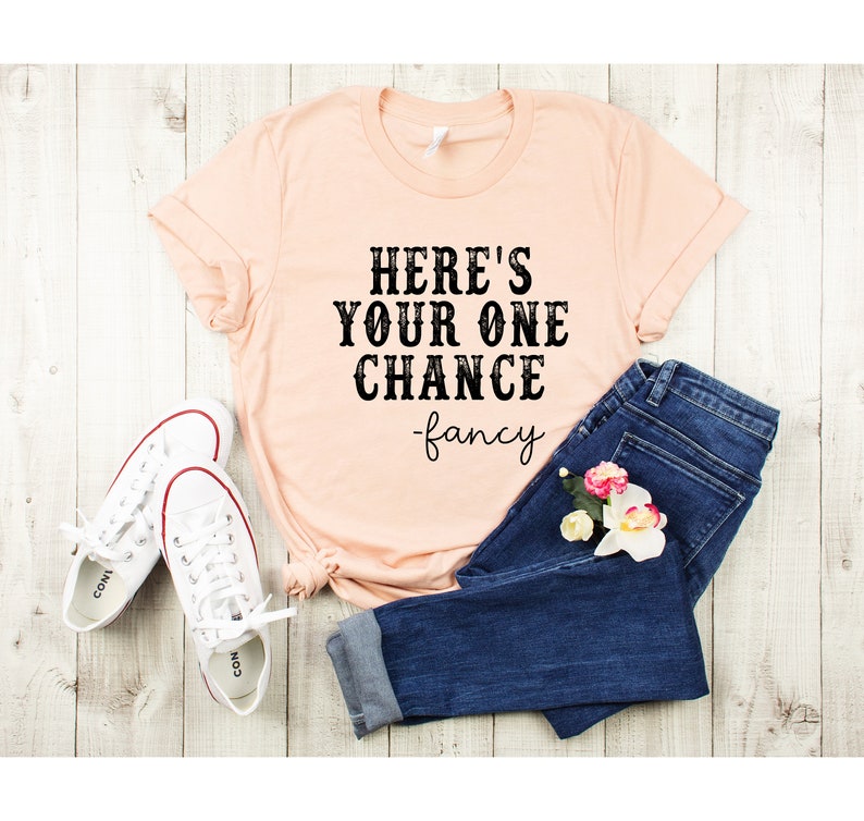 May include: A peach-colored t-shirt with the text "HERE'S YOUR ONE CHANCE -fancy" printed in black. The shirt is paired with blue jeans and white sneakers.