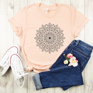 May include: A peach-colored t-shirt with a black mandala design. The t-shirt is paired with blue jeans and white sneakers.