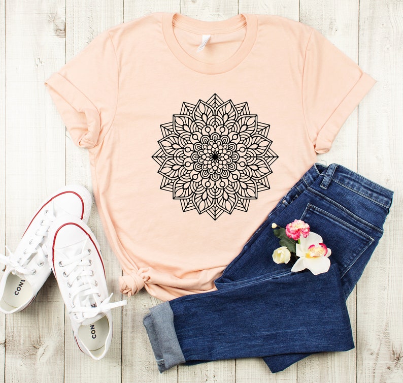 May include: A peach-colored t-shirt with a black mandala design. The t-shirt is paired with blue jeans and white sneakers.