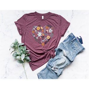 K&ouml;nnte beinhalten: Ein maroonfarbenes T-Shirt mit einem floralen Herz-Design. Das Herz besteht aus verschiedenen Blumen in Blaut&ouml;nen, Gelb und Gr&uuml;n. Das Shirt wird auf einer wei&szlig;en Marmorfl&auml;che mit einem Zweig Gr&uuml;n und einer blauen Jeans gezeigt.